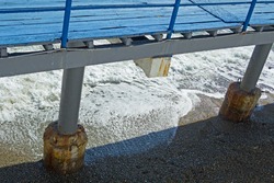 An old wharf covered with blue painted planks. Below are the concrete pillars of the pier. There is a pebble beach under the pier. The waves of the surf roll on the beach. 