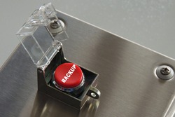 Round, red button with safety hood raised. The button is located on a metal box and is ready to use. The button has the inscription 