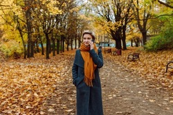 A blonde in a coat and scarf talks on the phone as she walks through an autumn park
