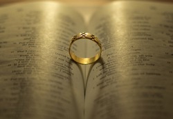 God Jesus Christ's blessing on the marriage relationship between husband and wife. Shiny golden engagement ring on open Holy Bible shadow forming heart. Faithful love, commitment, devotion, covenant.
