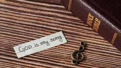 A rustic old treble clef note, handwritten quote: God is my song, and closed Holy Bible on wooden table. Christian biblical concept of music, praise, worship, and joy in the LORD. A closeup.