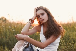 Boho lifestyle. Fashion portrait of beautiful young pretty girl with hippie outfit outdoors in the field at sunset. Soft warm vintage color tone. Artsy Bohemian Style. Horizontal. Eye contact