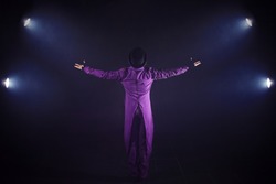 Young man in purple suit standing on the background of the spotlight. Showman spreading hands, show begins. Back view
