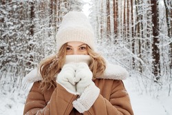 Winter walk, a young beautiful blonde in winter clothes walking in a snowy forest, a beautiful frosty day. A young woman warms her nose wrapped in a white scarf