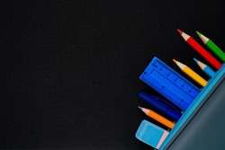 Blackboard with school stationery in pencil case, space for text. Bright colored pencils, pen, ruler, drawing pencil and eraser in the corner with space for promotional text on a black background.