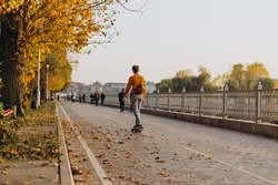 Man on skateboard on autumn promenade. A guy in yellow sweater with headphones rides a skateboard in alley on warm autumn day. Athletic man on city embankment of lake on a background of yellow leaves.