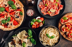 Full table of italian meals on plates Pizza, pasta, ravioli, carpaccio. caprese salad and tomato bruschetta on black background. Top view