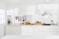 Modern white kitchen with counter and white details, minimalist interior with sunlight in daytime. Full set of kitchen equipment, pan, pot, electric hob, flipper, vegetable, fruit.
