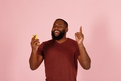 Cheerful young African-American man in t-shirt with mobile phone gets genius idea on light pink background in studio closeup