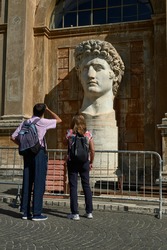 two tourists contemplate the head of the emperor Caesar Octavian Augustus on a pedestal illuminated from the side by the sun