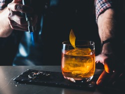 Bright orange Old Fashioned Cocktail with whiskey on a black board. Decorated with burning coal with the smoke. Bartender hand burning the cocktail decoration. Perfect Serve example. Horizontal