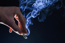 Male hand hold cigarette butt with smoke swirls dark background, nicotine addiction