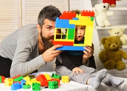 Family and childhood concept. Boy and bearded man play together on nursery background. Dad and kid hide behind plastic blocks wall. Father and son make grimaces looking through window of toy house