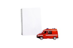 A toy red ambulance or other emergency vehicle next to a notebook. Isolated on white background. Place for your text.