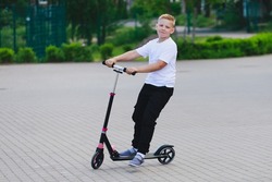 a blonde boy in a white T-shirt and black pants riding a scooter. High quality photo