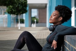 A young fit black woman is resting sitting on the floor and leaning on a big step. She is wearing big headphones around her neck and her eyes are closed. She looks relaxed.