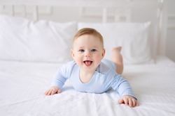 a cute healthy little baby is lying on a bed on white bedding at home in a blue bodysuit. The kid looks at the camera, smiles