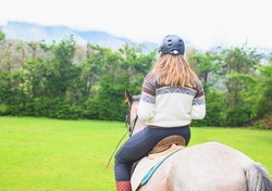 Back view of a caucasian woman wearing an andean sweater and riding a horse in the meadow. Horizontal. With copy space.