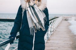 Young woman stands on the winter promenade against the sea, dressed in a down jacket, hood, gloves and warm scarf. Walk in cold season in warm clothes. Pier and seafront with snow on the background.