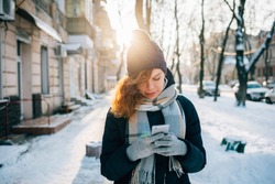 Young woman wearing warm scarf and knitted hat smiles and using mobile phone writing text message outdoors on sunny winter day in city.