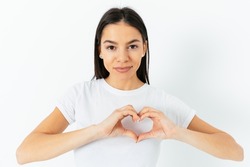 Young brunette woman showing heart symbol with fingers holding hands near chest, standing on white studio background