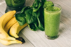 Vitamin Smoothies in a glass on a wooden table. On the table are also banana, spinach and a food processor.