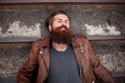 A drunken bearded rock guitarist with gray hair and a large beard in a brown leather jacket lies and smokes a cigarette on the old dirty steps