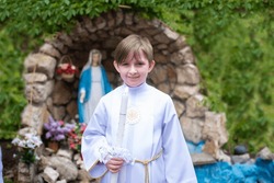 a child in white traditional festive clothes with a saint before his first communion stands in front of a statue of the virgin mary