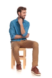 pensive young casual man looks to side while sitting on wooden chair on white background