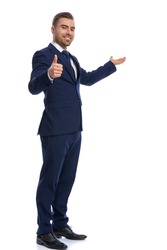 smiling unshaved guy in elegant suit making thumbs up sign and presenting to side, smiling and standing in a side view position isolated on white background, full body