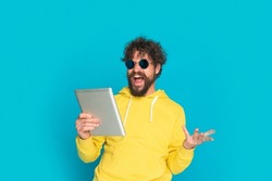 happy unshaved guy with curly hair and moustache wearing retro sunglasses and yellow hoodie, holding tab, making a hand gesture and laughing on blue background in studio