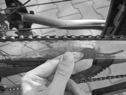 Greyscaled image - Close view on manual checking of bike chain wear and tear degree by measuring the length of segments with a transparent plastic ruler.