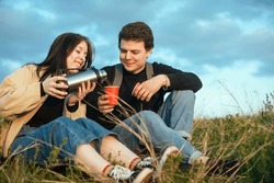 a couple of tourists in love a guy and a girl during outdoor recreation sit on the ground and drink a hot drink of coffee, tea from a thermos