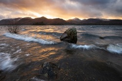 Crashing waves on lake shoreline with dramatic sunset sky above mountains at Derwentwater in the Lake District, UK.