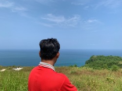 a man with a red shirt standing on the hill looking at the beautiful scenery of the open sea in Pacitan, Indonesia.