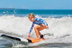 Happy baby girl - young surfer ride on surfboard with fun on sea waves. Active family lifestyle, kids outdoor water sport lessons and swimming activity in surf camp. Summer vacation with child.