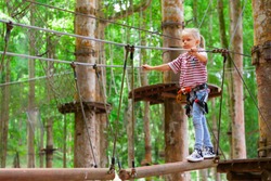 Brave little child in safety harness climb on tree tops route to zip line, pass hanging bridge obstacle in adventure rope park. Outdoor activity, family lifestyle in summer camp on vacation with kid
