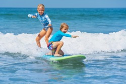 Happy baby boy and girl - young surfers ride with fun on one surfboard. Active family lifestyle, kids outdoor water sport lessons, swimming activity in surf camp. Sea beach summer holiday with child.