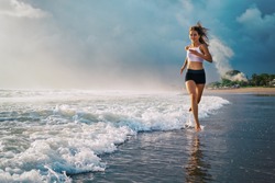 Active sporty woman run along ocean surf by water pool to keep fit and health. Sunset black sand beach background with sun. Woman fitness, jogging training and sport activity on summer family holiday.