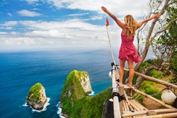Family vacation lifestyle. Happy woman with raised in air hand stand at viewpoint. Look at Kelingking beach under high cliff. Travel destination in Bali. Popular place to visit on Nusa Penida island.