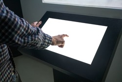 Mockup closeup image: man hand touching white empty interactive touchscreen display kiosk in dark room of technology museum. Mock up, template, scifi, education, futuristic and technology concept