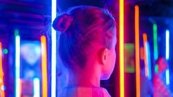 Back view - portrait of woman looking right at interactive exhibition or museum with colorful fluorescent tube illumination. Futuristic, retrowave, immersive, entertainment concept
