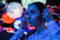 Woman wearing wireless black headphones and looking around in dark room of interactive exhibition or museum with colorful illumination. Futuristic, entertainment, immersive concept
