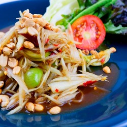 Papaya salad with traditional pickled fish cooked with papaya noodles, tomatoes, long beans and sprinkled with peanuts. Dried shrimps with a side of lettuce put in a blue dish.
