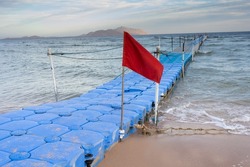 Red warning flag on beach. Blue plastic pontoon in sea water. Swimming forbidden in windy weather, ocean waves, storm