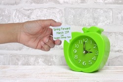 Spring Forward March 2022 Daylight Savings Time comment box held in held next to clock on whitewashed wood 
