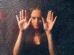 Quarantine self isolation. Pandemic anxiety. Social distancing. Textured art portrait of bored unhappy annoyed trapped woman in black touching plastic bubble wrap wall in darkness.
