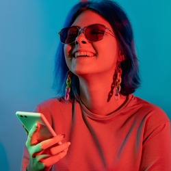Phone entertainment. Millennial lifestyle. Mobile technology. Joyful laughing woman using smartphone in red neon light isolated on blue background.