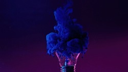 Ink water drop. Color smoke cloud. Fantasy imagination. Neon blue fluid in broken light bulb on purple gradient grain texture abstract art copy space background.