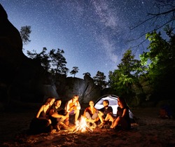 Happy Friends hikers having a rest together around campfire beside camp and illuminated tourist tent at night. On background beautiful night starry sky full of stars and Milky way, mountain rocks.